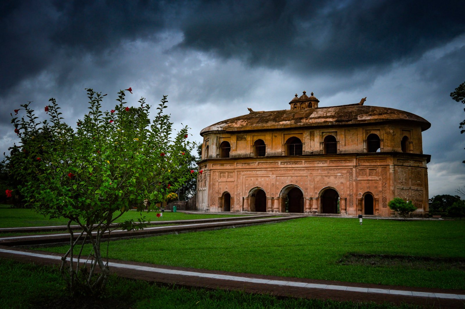 a building with a circular roof and a green lawn with a tree and a cloudy sky